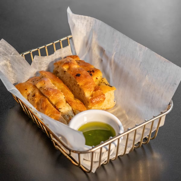 Golden focaccia bread slices in a wire basket with parchment paper and a small dish of olive oil.