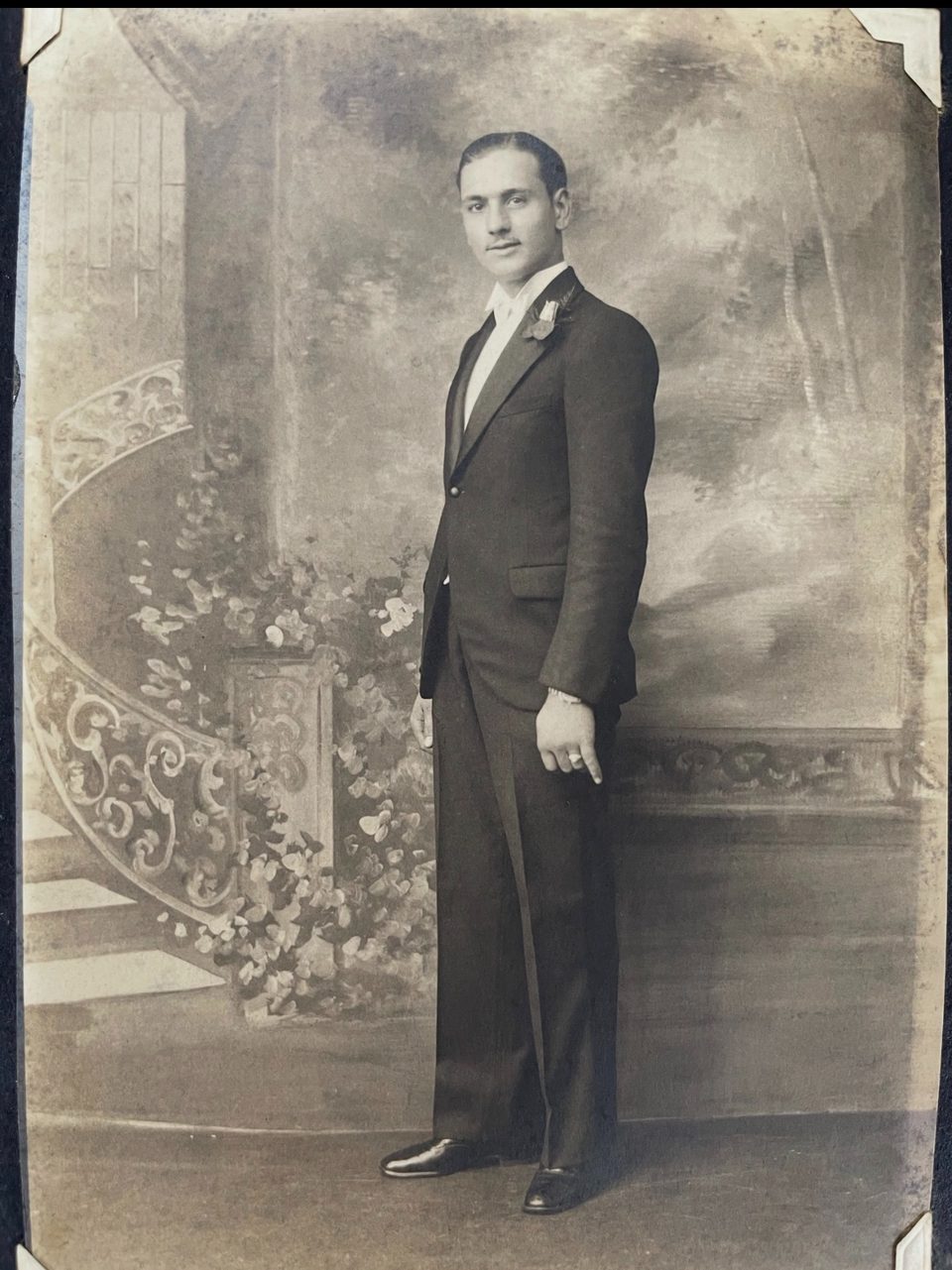 A vintage black and white portrait of a man in a tuxedo standing next to a painted staircase.