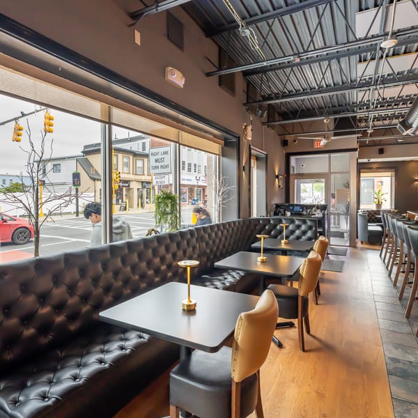 Modern restaurant interior with black tufted booths, small tables, and a long bar with stools.