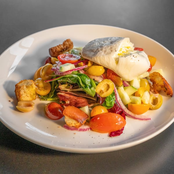 A vibrant salad with burrata cheese, cherry tomatoes, red onion, olives, and croutons on a white plate.