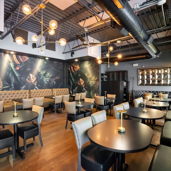 Modern restaurant interior with dark tables, light chairs, banquette seating, and a tropical leaf mural.
