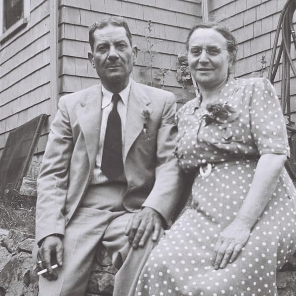 A black and white photo of a man and woman sitting outdoors, the man holding a cigarette.