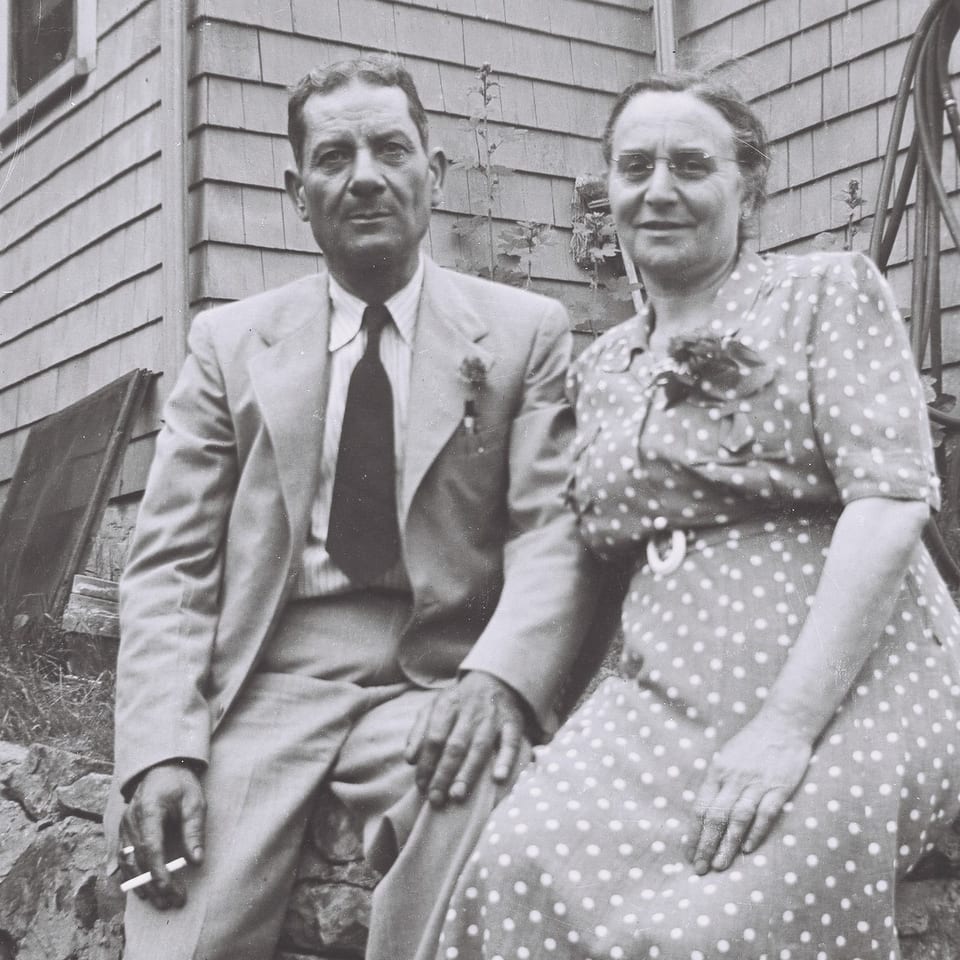 A black and white photo of a man and woman sitting outdoors, the man holding a cigarette.
