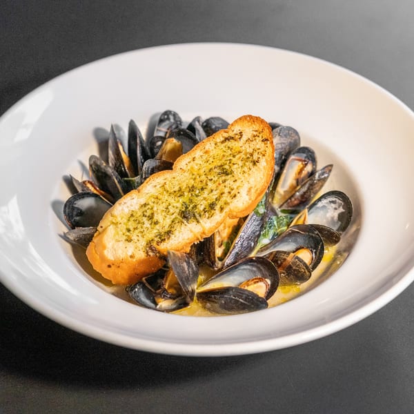 A white bowl filled with mussels in a creamy sauce, topped with a slice of garlic bread.