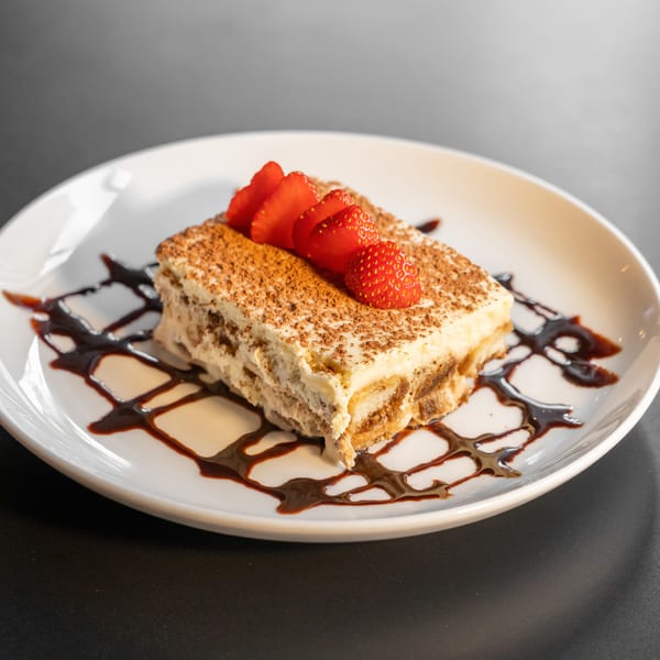 A slice of tiramisu with cocoa powder, fresh strawberries, and chocolate drizzle on a white plate.